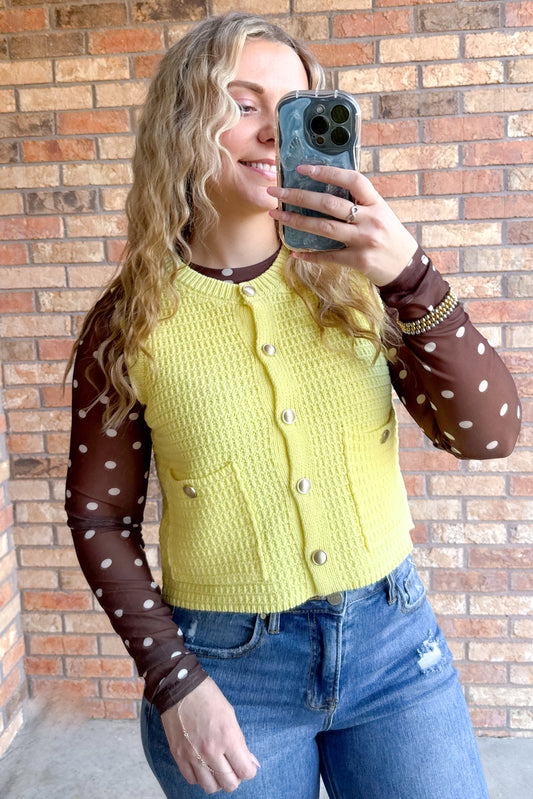 This textured sleeveless knit yellow vest features a ribbed crew neckline and a polished button-up front adorned with gold buttons. The fitted silhouette is accentuated by two front patch pockets. Cut to a waist-level cropped length, this piece can be worn open or closed.