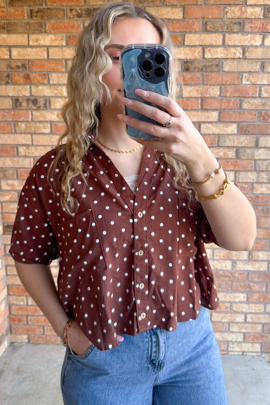A relaxed fit button-up shirt with polka dot pattern and short sleeves. Features a classic collar and front pockets. Cropped. Chestnut colored top with small white polka dots. 