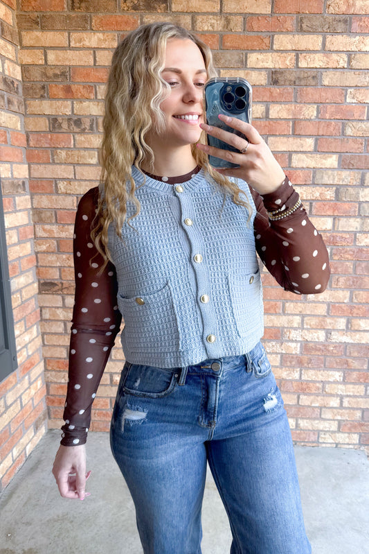 This textured sleeveless knit ice blue vest features a ribbed crew neckline and a polished button-up front adorned with gold buttons. The fitted silhouette is accentuated by two front patch pockets. Cut to a waist-level cropped length, this piece can be worn open or closed.